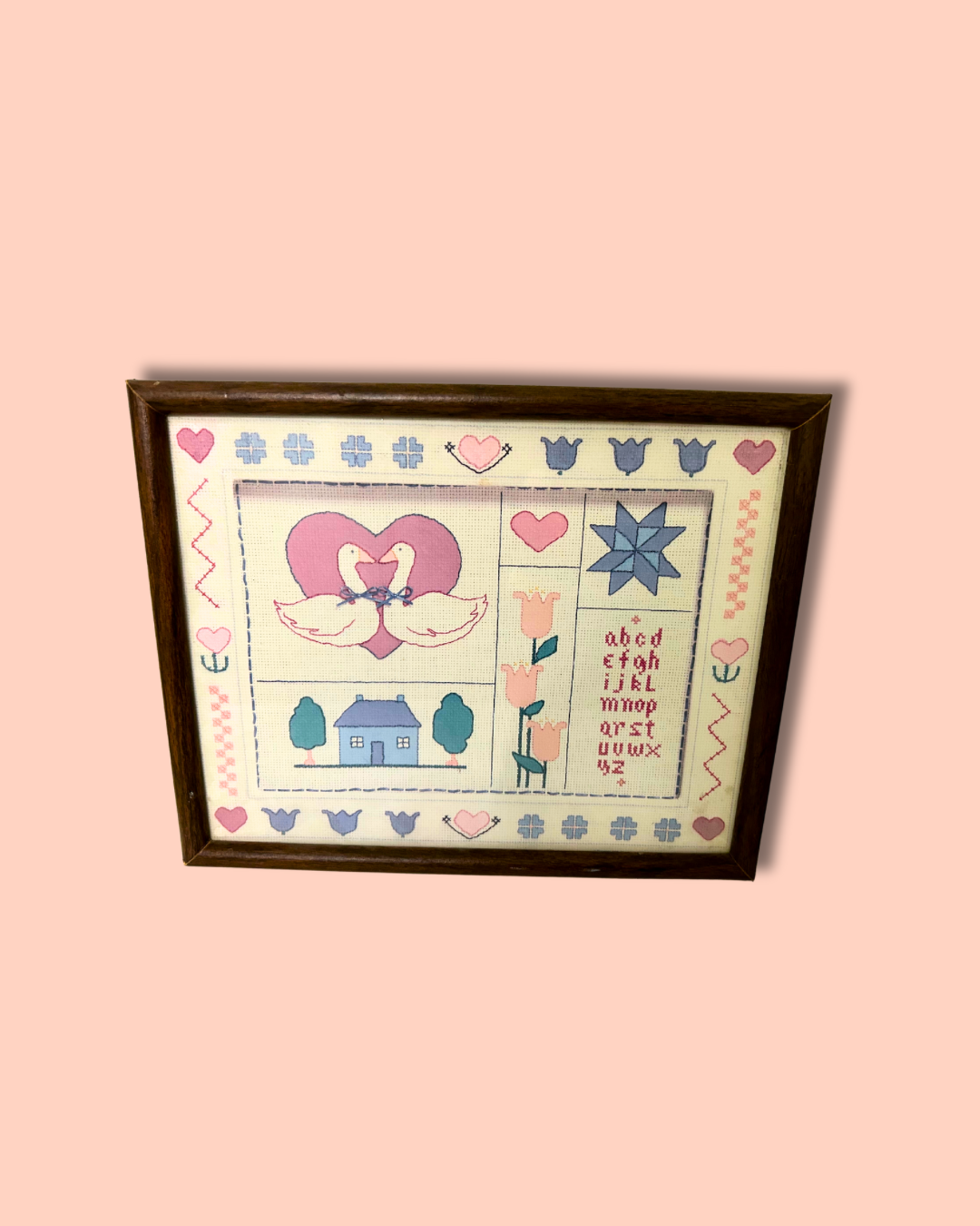 Framed cross-stitched piece with decorative elements on a pink background