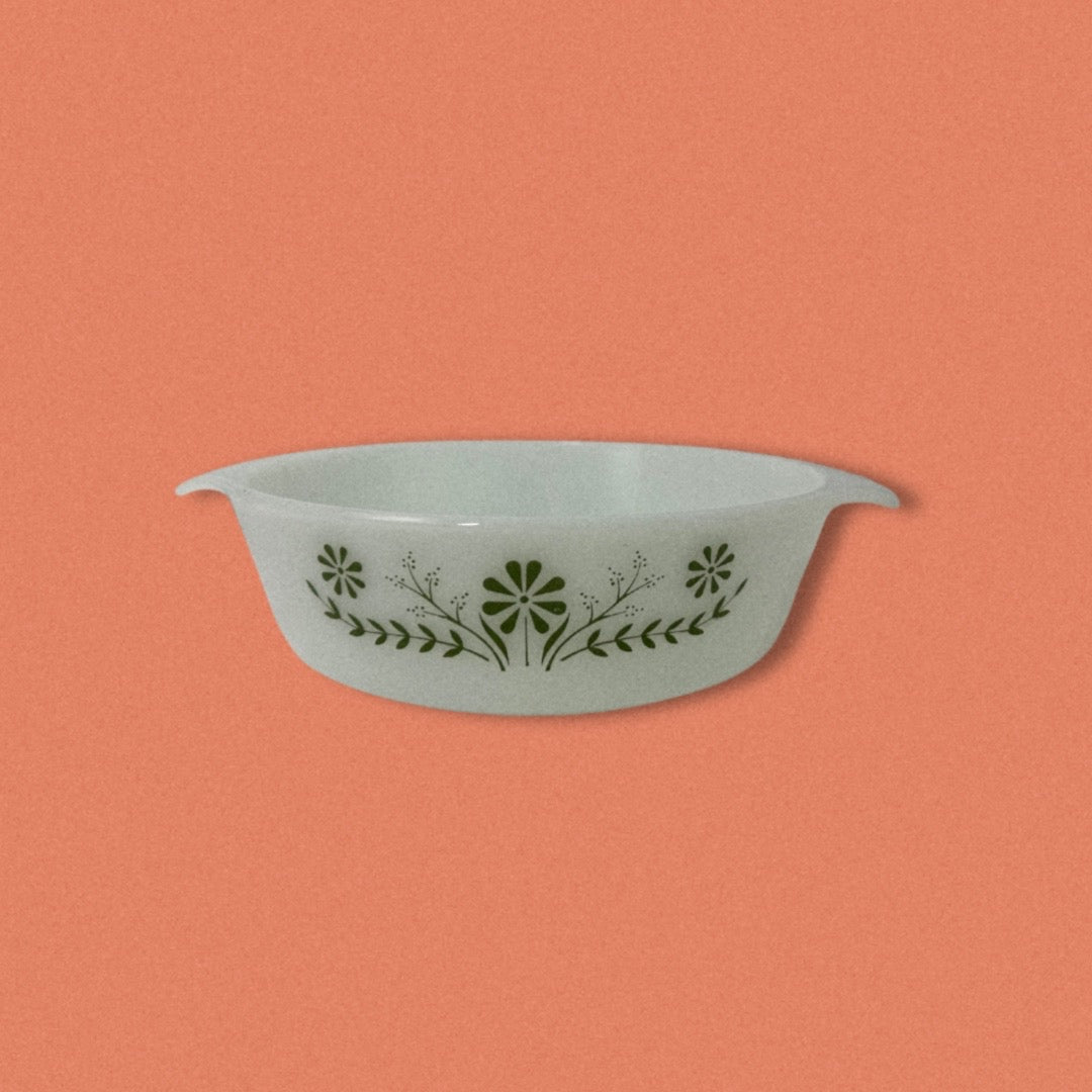 A vintage Glasbake green baking dish with a daisy pattern on the front, displayed against an orange background.