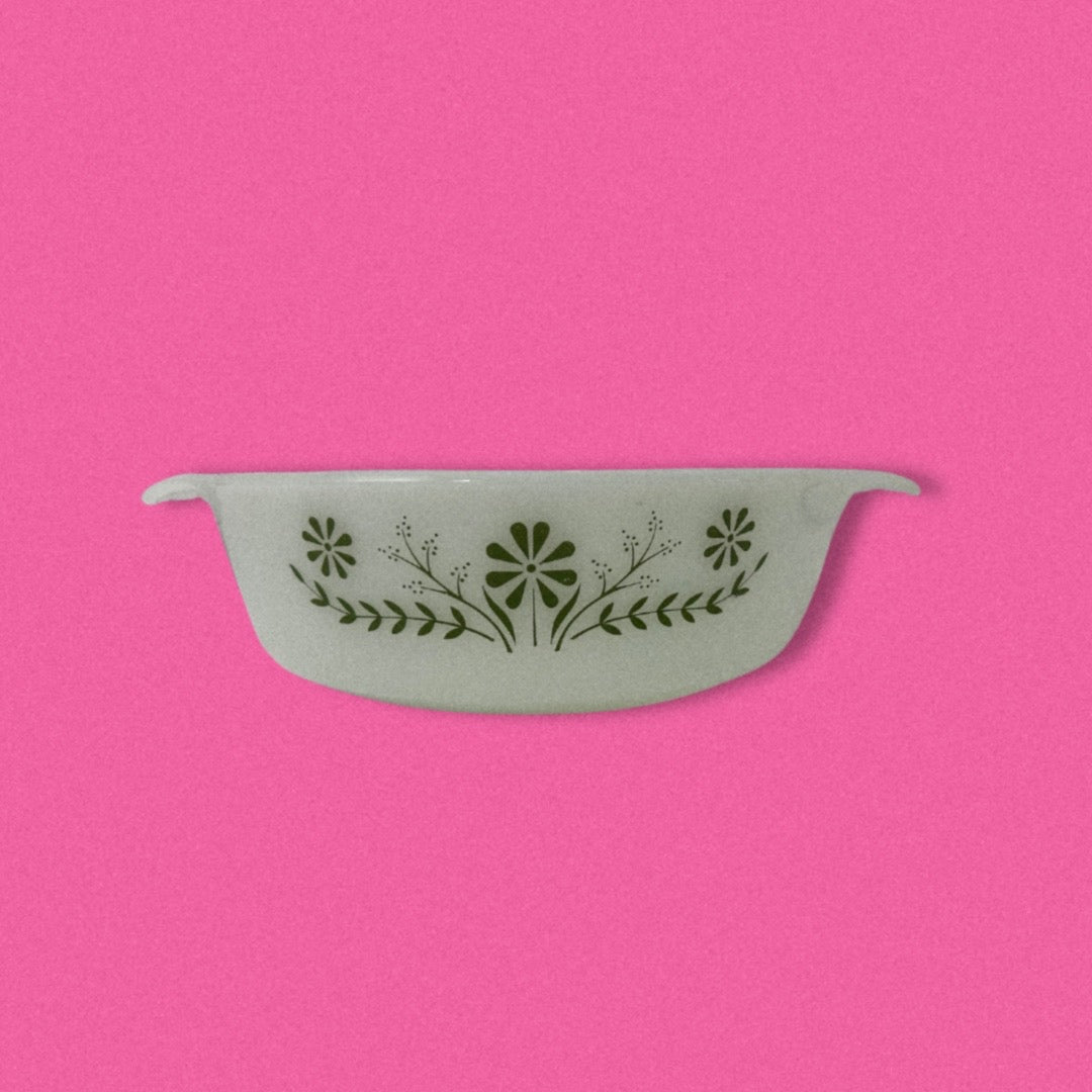 A vintage Glasbake green baking dish with a daisy pattern on the front, displayed against an pink background.
