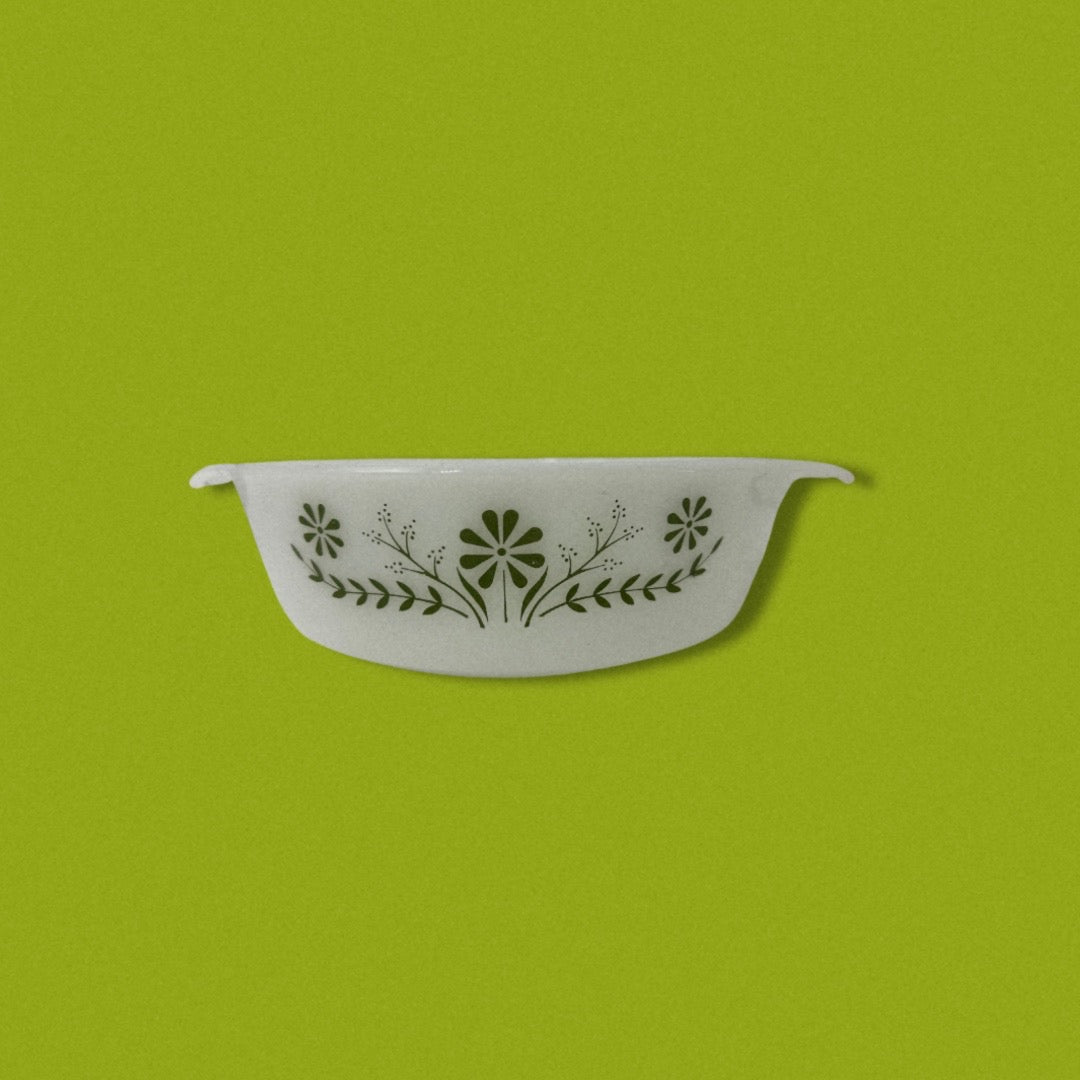 A vintage Glasbake green baking dish with a daisy pattern on the front, displayed against a green background.