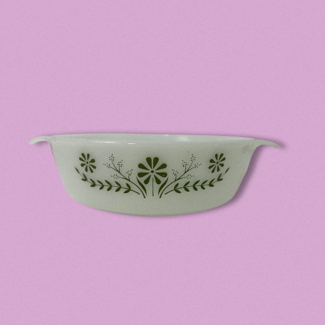 A vintage Glasbake green baking dish with a daisy pattern on the front, displayed against a purple background.