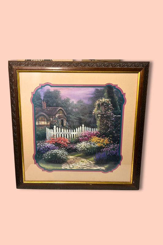 Framed artwork of a garden scene with a white picket fence on a pink background