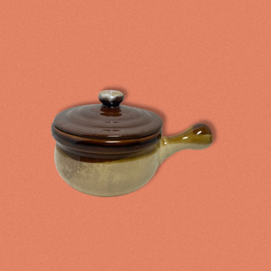A small two-tone mini stoneware pot with a brown lid and beige body, isolated on a reddish-pink background.