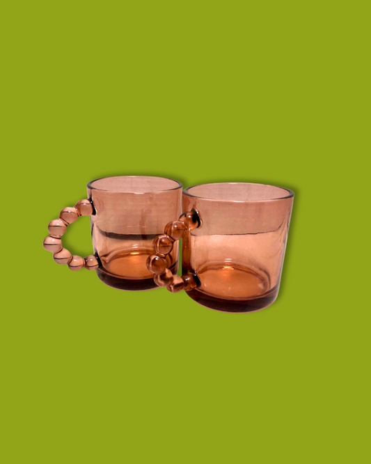 A pair of pink glass mugs with beaded handles on a green background.