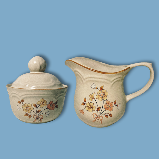 A vintage stoneware floral sugar jar with a lid and a creamer, both featuring hand-painted florals on a blue background.